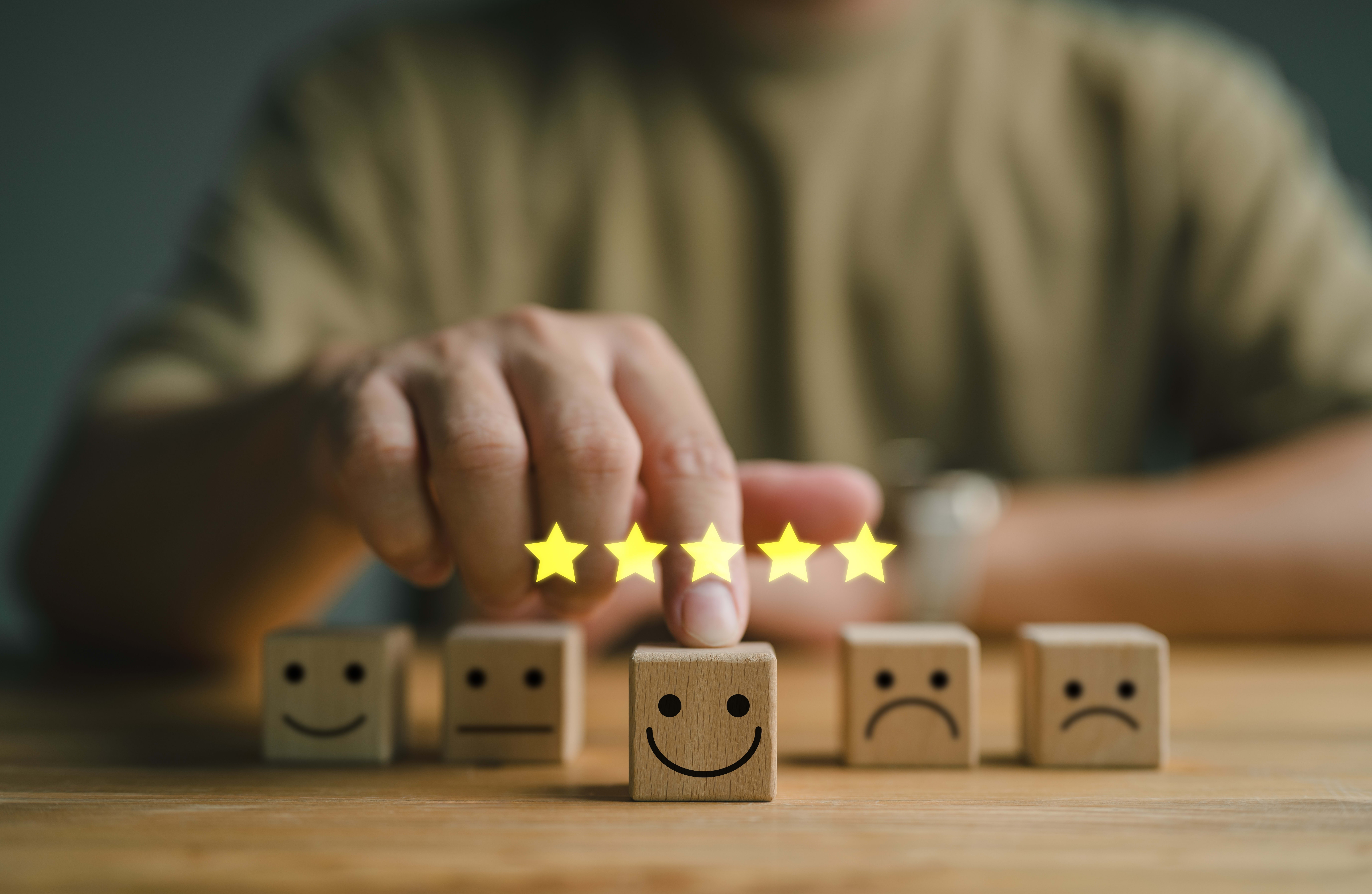 Hand pointing at a wooden block with a smiley face, surrounded by five glowing stars, representing a positive rating