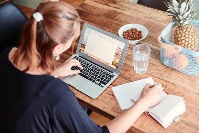 Woman at dining table with a computer, nuts, and fruits, writing on a notebook
