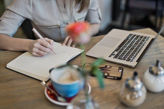 Person sitting on a table, writing in a notebook with a cup of coffee, a phone, and a laptop