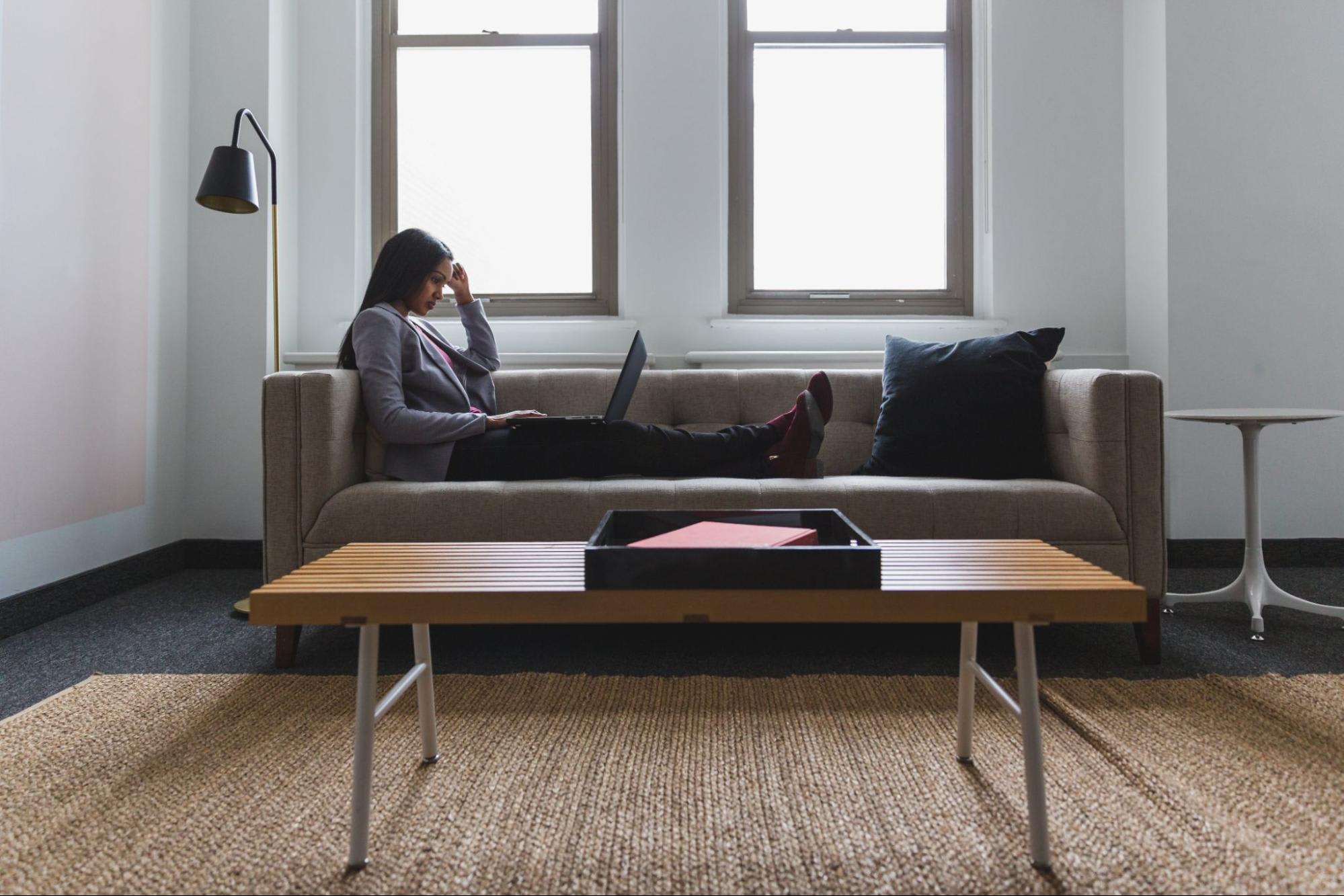 A woman sits working with her legs up on the couch and a laptop computer in her lap.