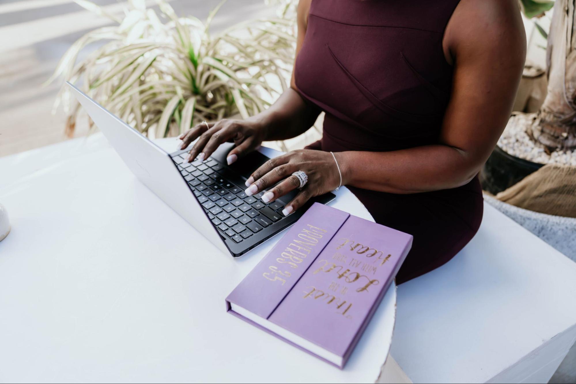 Person sits at a white table with a laptop on front of them, a purple notebook beside them and a large plant behind them.